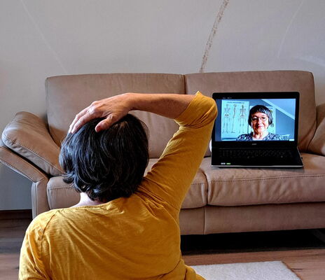 Eine Frau macht eine Feldenkrais-Übung. Sie sitzt auf dem Boden mit dem Rücken zur Kamera. Ihre rechte Hand ist auf dem Kopf und sie schaut auf einen Laptop, der vor ihr auf einem Ledersofa steht.