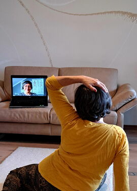 Eine Frau macht eine Feldenkrais-Übung. Sie sitzt auf dem Boden mit dem Rücken zur Kamera. Ihre linke Hand ist auf dem Kopf und sie schaut auf einen Laptop, der vor ihr auf einem Ledersofa steht.