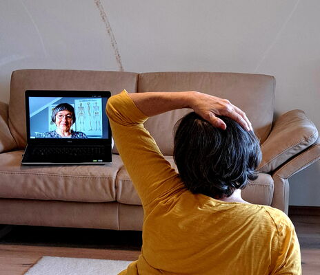 Eine Frau macht eine Feldenkrais-Übung. Sie sitzt auf dem Boden mit dem Rücken zur Kamera. Ihre linke Hand ist auf dem Kopf und sie schaut auf einen Laptop, der vor ihr auf einem Ledersofa steht.