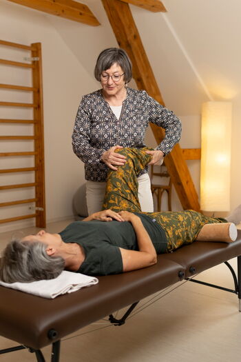 Eine Situation aus einer Einzelstunde in Feldenkrais Funktionale Integration. Eine Person liegt auf einer Massageliege. Die Feldenkrais-Pädagogin steht neben der Liege und hält das linke Bein der Liegenden.