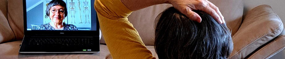 Eine Frau macht eine Feldenkrais-Übung. Sie sitzt auf dem Boden mit dem Rücken zur Kamera. Ihre linke Hand ist auf dem Kopf und sie schaut auf einen Laptop, der vor ihr auf einem Ledersofa steht.