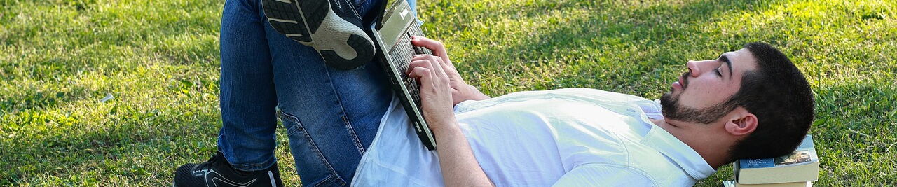 Ein Mann liegt auf einer Wiese. Sein Kopf ist auf einige Taschenbücher gestützt. Auf den aufgestellten Beinen hat er seinen Laptop, in den er gerade etwas tippt.